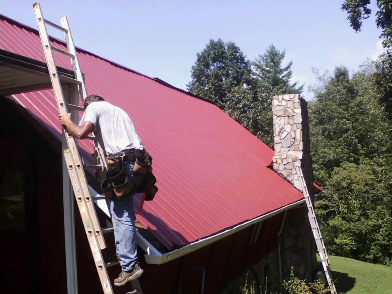 Roofer on a ladder beside a red metal roof and stone chimney — Soffit & Fascia Repair by Roofing Brothers in Middlebranch